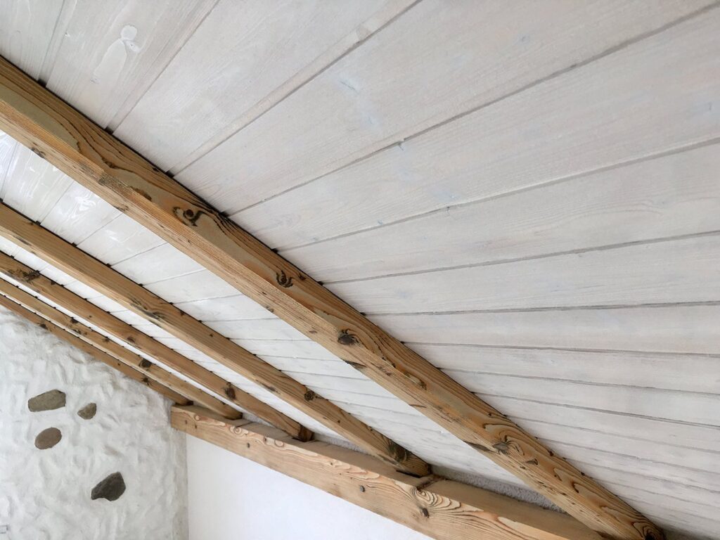 Weiss gestrichene Holzdecke mit hellen Sparren über einer verputzten Bruchsteinwand