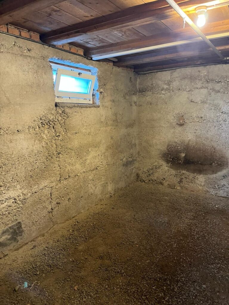 Unsanierter Kellerraum mit rohen Betonwänden, dunklen Feuchtflecken und kleinem Fenster