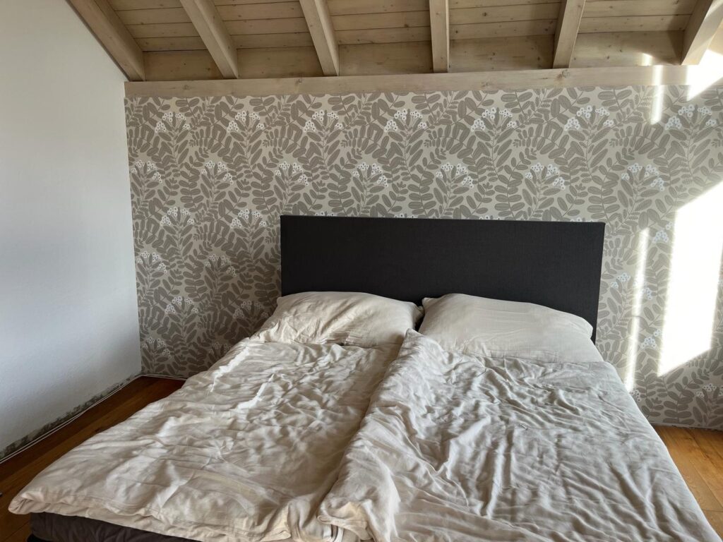 Schlafzimmer mit blattgemusterter Tapete hinter dem Bett unter einer hellen Holzbalkendecke