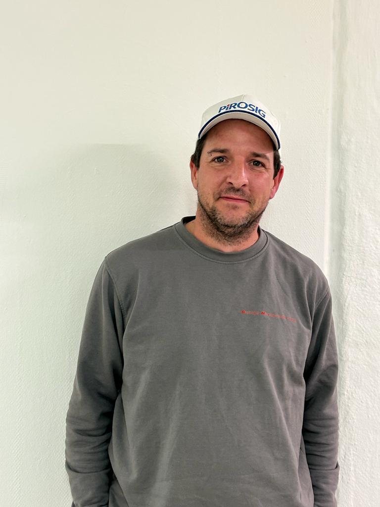 Florian Giger mit weisser Kappe und grauem Firmenpullover vor einer weissen Wand