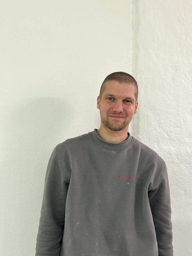 Stefan Gisler im grauen Arbeitspullover vor einer weissen Wand