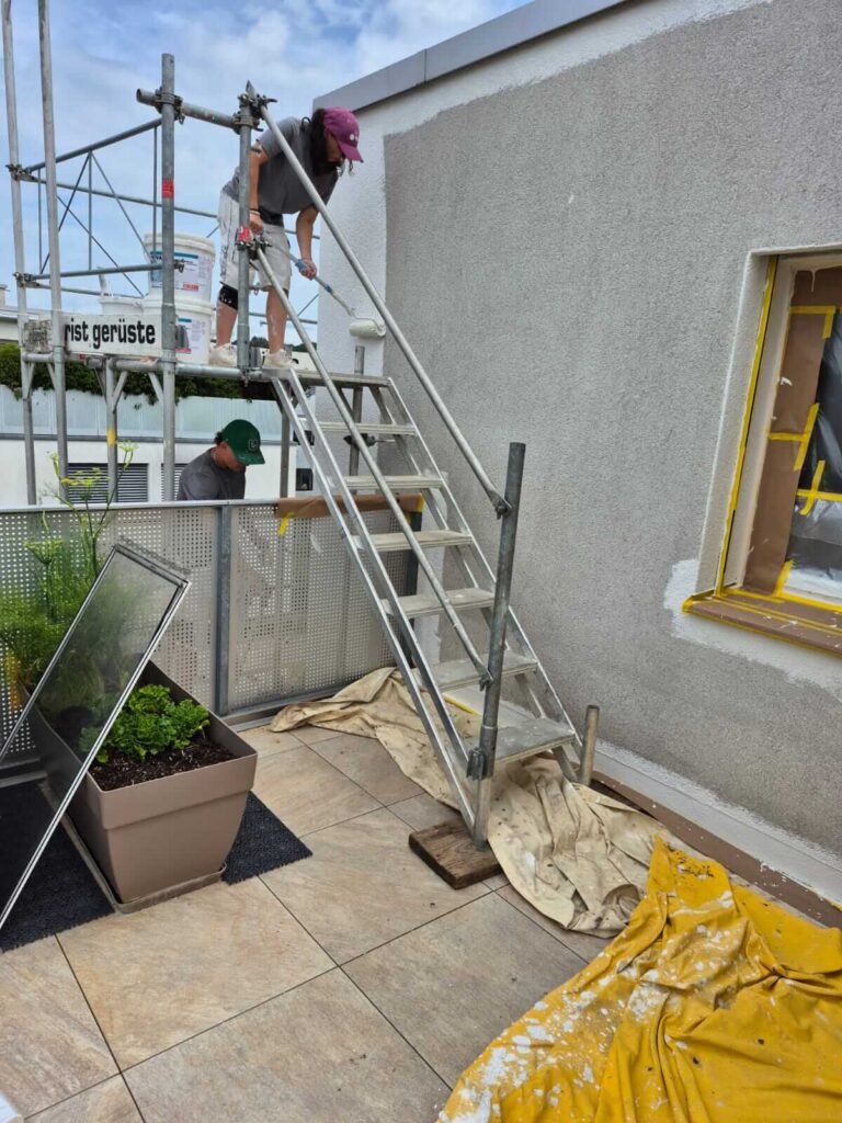 Zwei Maler streichen von einem Gerüst aus die hellgraue Fassade eines Hauses, die Fenster sind sorgfältig abgeklebt