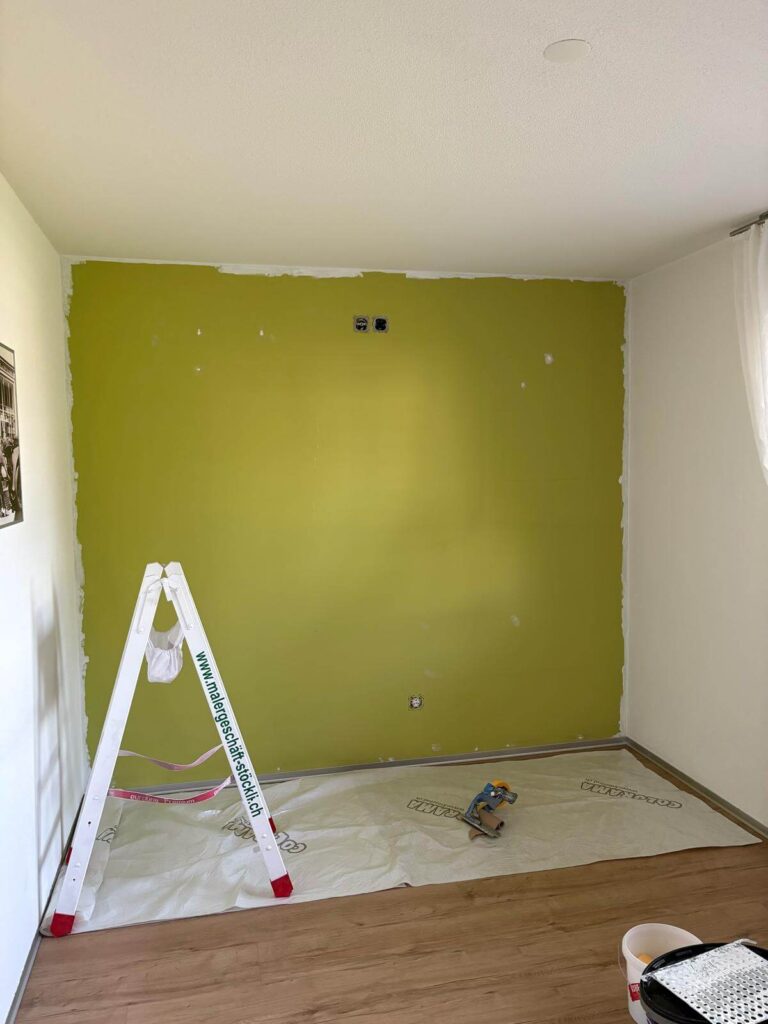 Wohnzimmerwand in Olivgrün mit ausgebesserten Rändern, davor eine Malerleiter auf Abdeckvlies