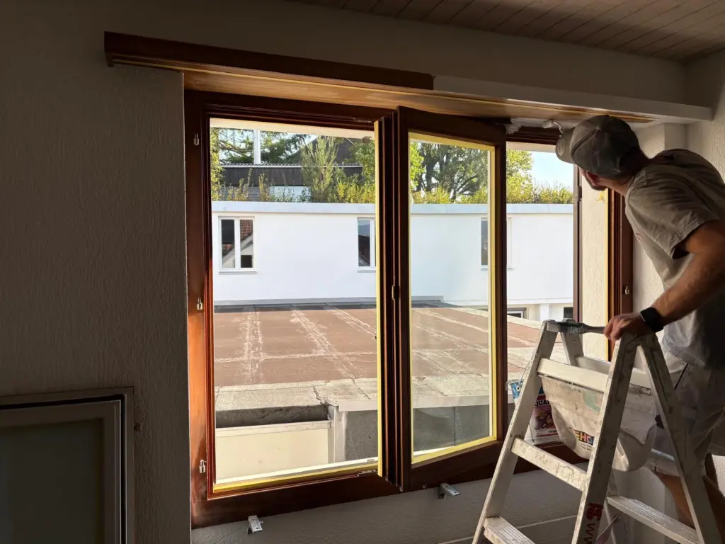 Maler auf der Leiter lackiert den Holzrahmen über einem grossen Fenster