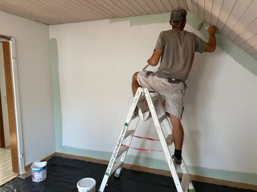 Maler auf der Leiter streicht eine Wand unter der Holzdecke in hellem Mintgrün, der Boden ist abgedeckt